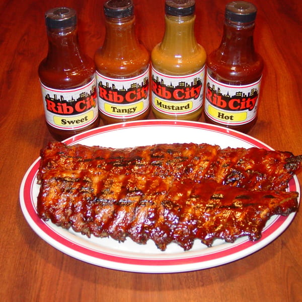 A rack of Rib City BBQ ribs on a plate with four bottles of Rib City sauces.