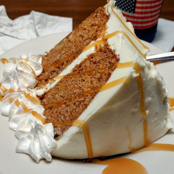 A slice of carrot cake with white frosting, caramel drizzle, and whipped cream on a white plate.